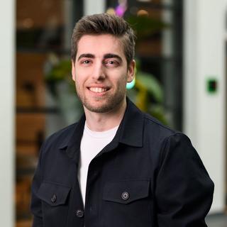 a man in a black jacket smiling while standing in front of a building